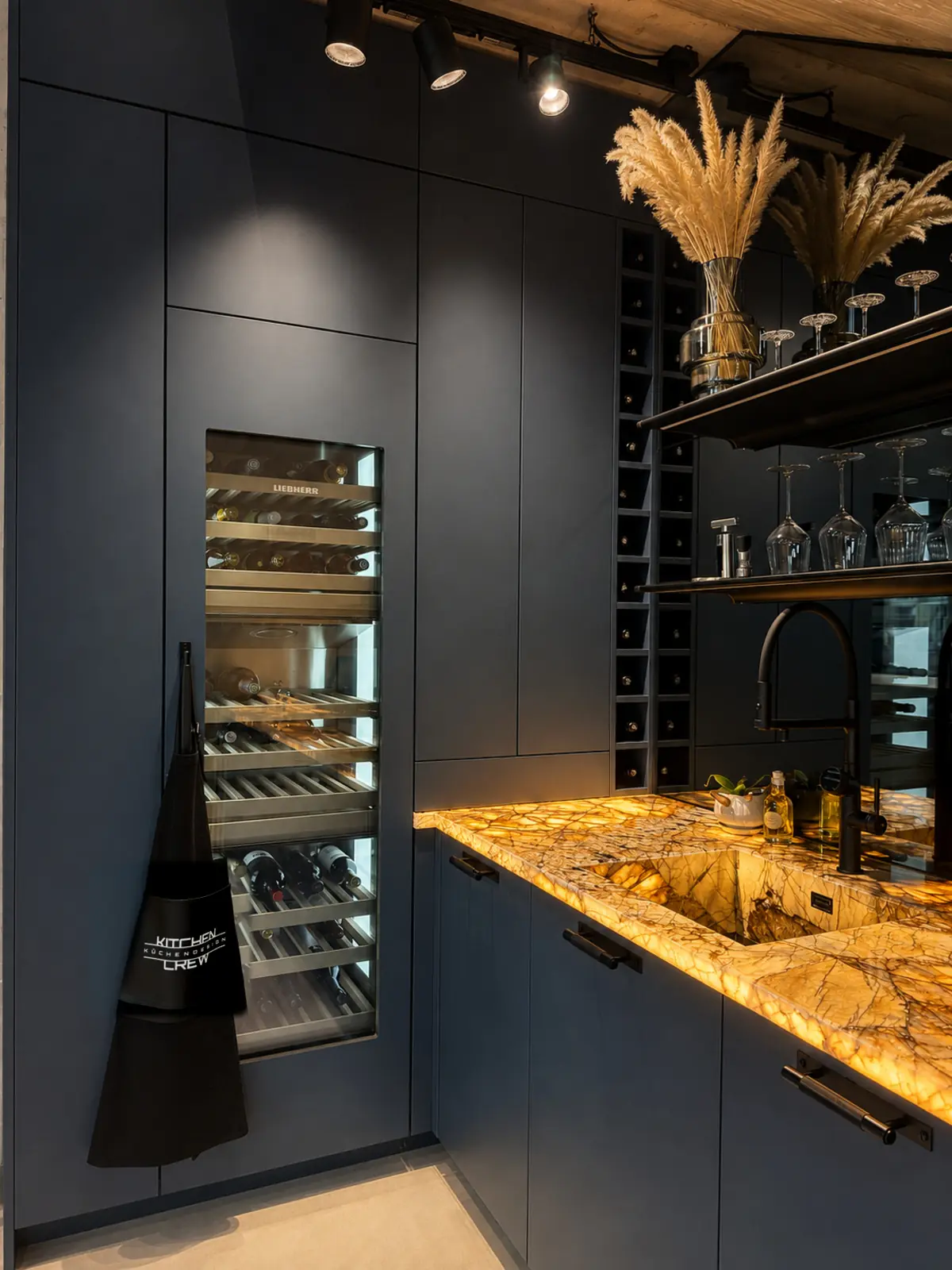 Beleuchtete goldene Naturstein-Theke mit Spüle und schwarzer Armatur neben Weinkühlschrank und Weinregal